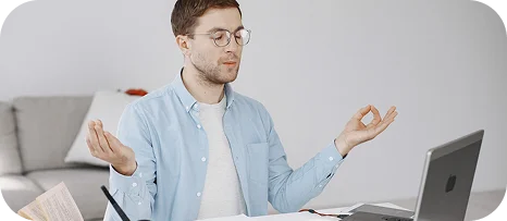 Jonge man die aan zijn bureau mediteert terwijl hij op een laptop studeert, waarbij hij zich concentreert en stress beheerst tijdens het online leren.